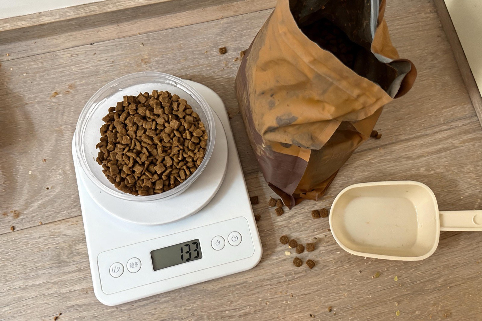 A digital kitchen scale on a counter with kibble in a plastic container, reading 133 grams.